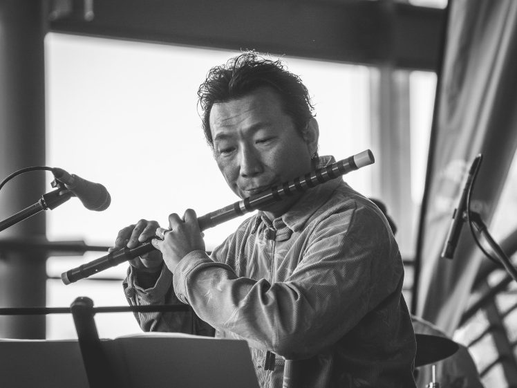 Image of A musician plays a flute in front of a microphone during a performance. He appears focused, and the image is in black and white with a blurred background.