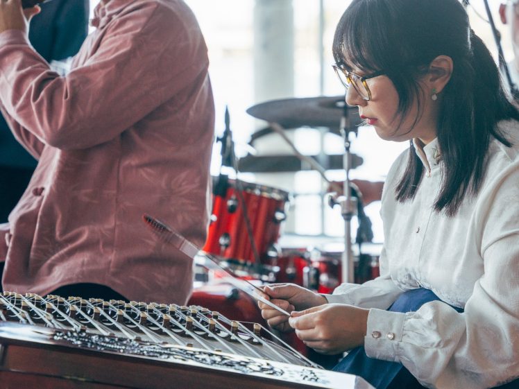 Image of A woman in spectacles plays a traditional stringed instrument with mallets, whilst other musicians perform in the background with drums and wind instruments. The scene appears to be part of a live music performance.