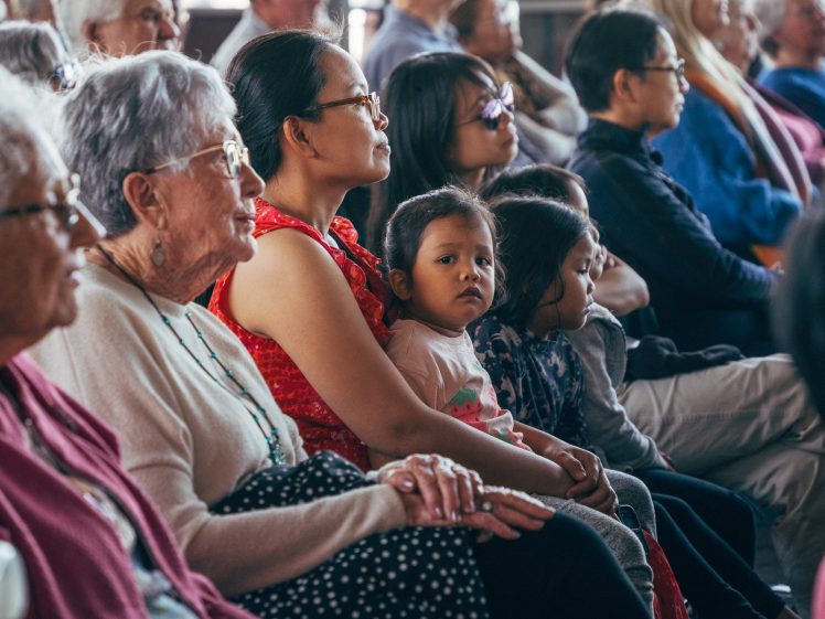 Image of A diverse group of people of various ages sit closely together in rows, attentively watching something off-camera. A young child in the centre looks directly at the camera whilst others face forward.