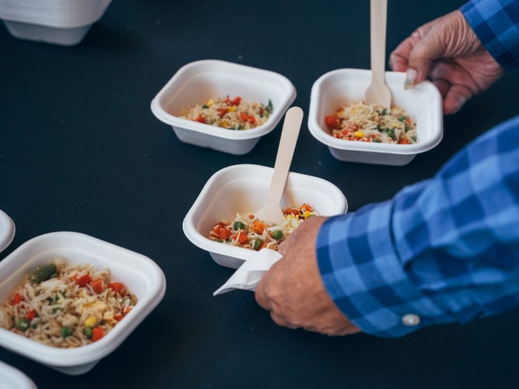 Image of A person in a blue checked shirt places disposable forks into small containers of mixed rice and vegetables on a black table.