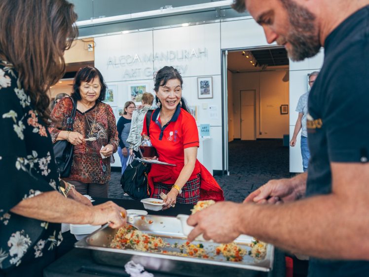 Image of People are serving food from trays at a communal table indoors. A smiling woman in a red shirt is receiving food, whilst others wait and watch. Artworks hang on the wall in the background.