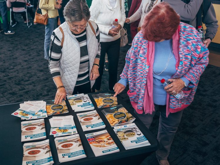 Image of Two older women stand at a table covered with leaflets and magazines, looking through them. One woman holds a water bottle, and other people are visible in the background.