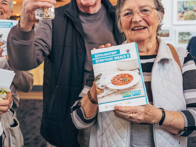 Image of Two smiling older adults stand indoors. One holds a bottle of water, while the other holds a LiveLighter Everyday Meals booklet. They appear cheerful and are promoting healthy living materials.