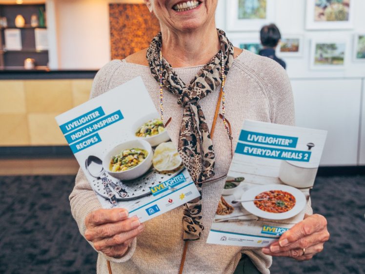 Image of An older woman with short grey hair smiles whilst holding two recipe booklets, titled Indian-Inspired Recipes and Everyday Meals 2, in a brightly lit gallery space.