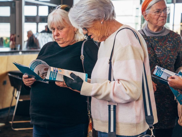 Image of Three older women stand indoors, reading leaflets. They appear focused and engaged, wearing casual clothing and spectacles. The background shows windows and a modern interior.