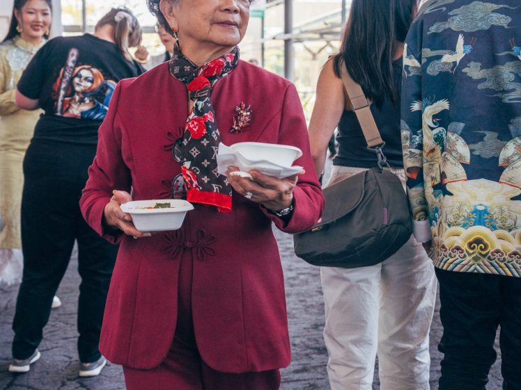 Image of An elderly woman in a red suit carries food containers at an indoor event, surrounded by people walking and talking in the background. Bright natural light comes through large windows.