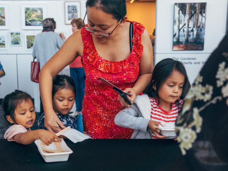 Image of A woman in a red dress helps three young girls at a table, handing them items from a container. Artworks are displayed on the wall behind them in a gallery setting.