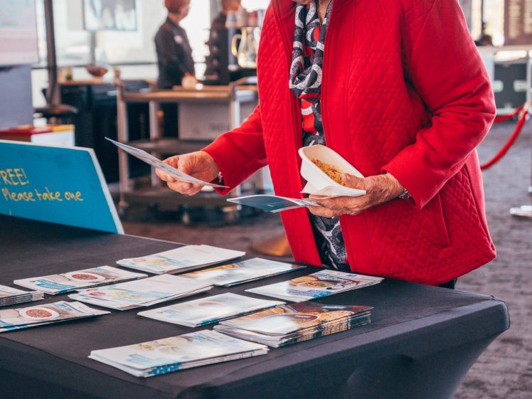 Image of An older woman in a red jacket and sunglasses examines brochures on a black table in a bright, modern indoor space. She holds food in one hand and looks attentively at the papers.