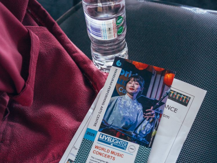 Image of A plastic water bottle, a maroon fabric, and concert brochures rest on a grey chair. The brochures display images and text about world music concerts.