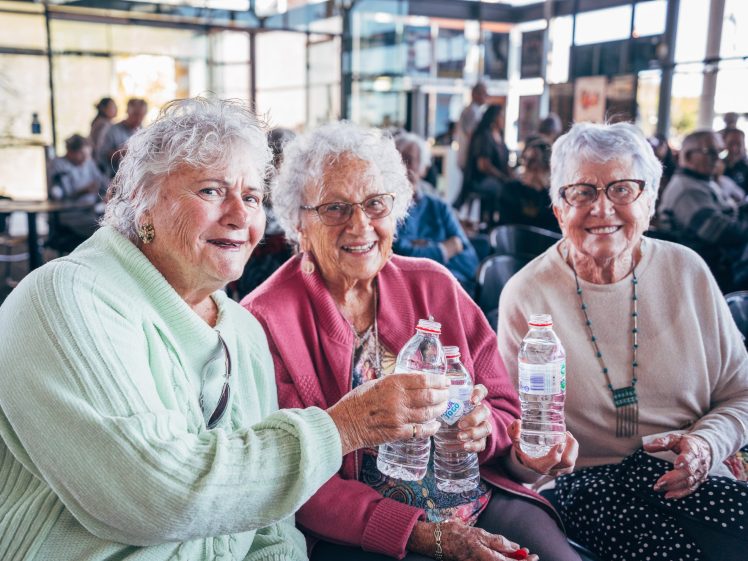 Image of Three elderly women sit closely together indoors, smiling and holding bottled water. They are dressed in colourful jumpers and spectacles, with other people visible in the background. The atmosphere appears cheerful and sociable.