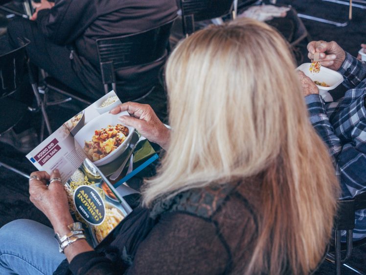 Image of A person with long blonde hair reads a food magazine featuring a bowl of pasta whilst another person next to them eats from a small bowl with a spoon in a seated, indoor setting.