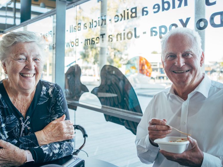 Image of An older woman and man sit at a table in a bright café, smiling. The man holds a small white bowl and spoon, while the woman has her hands folded on the table. Large windows and sunlight are visible in the background.