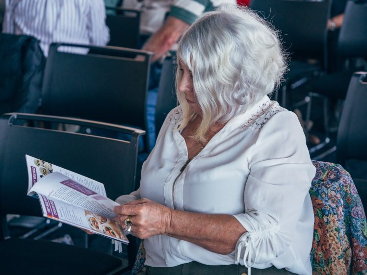 Image of An older woman with white hair wearing a white blouse sits in a chair and reads a leaflet in a room with several empty chairs around her.