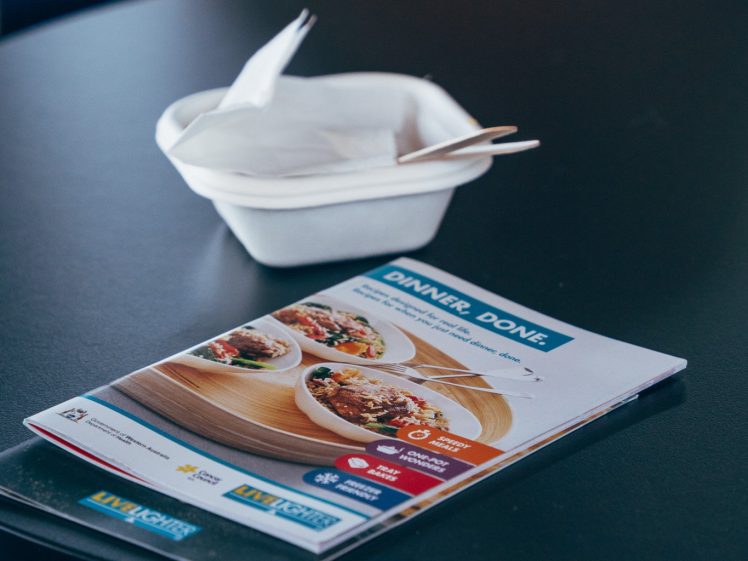 Image of A dinner menu with images of food dishes lies on a dark round table next to a white plate holding folded serviettes and cutlery.