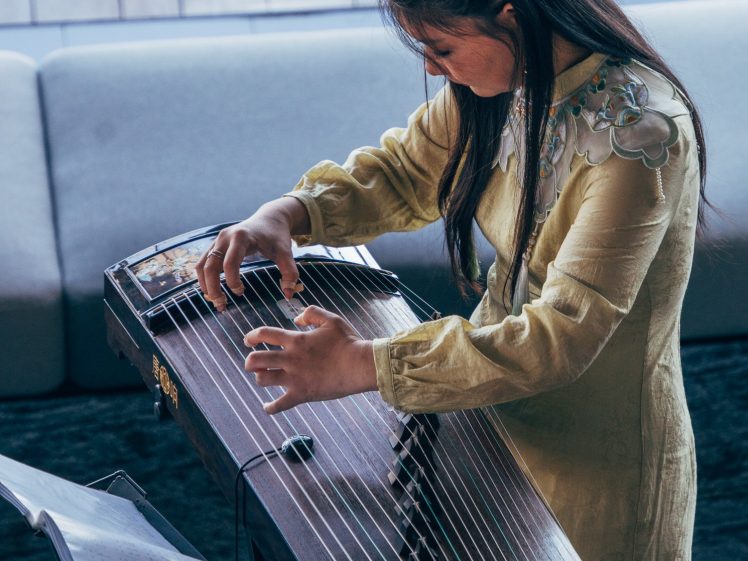 Image of A woman in a yellow traditional dress plays a guzheng, a Chinese string instrument. She is focused on the strings, with sheet music on a stand beside her. The background shows a sofa and a wooden floor.