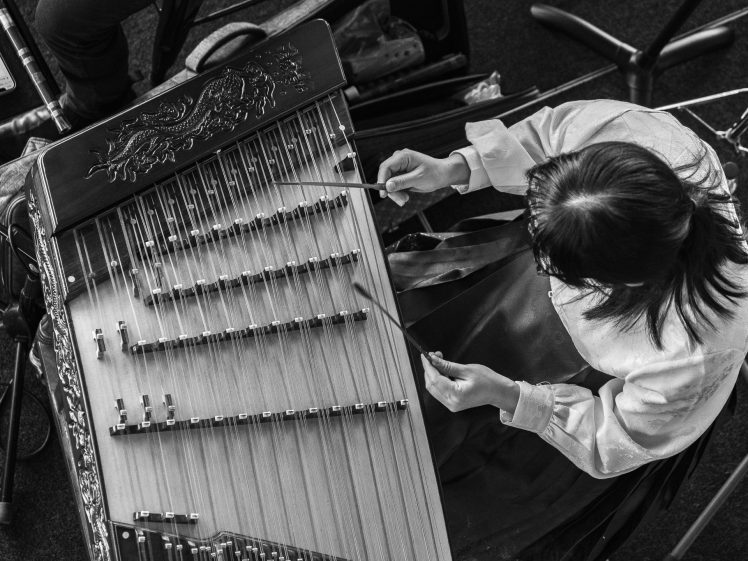 Image of A person in traditional clothing plays a stringed musical instrument with mallets, viewed from above in black and white. The instrument has ornate carvings and multiple strings.