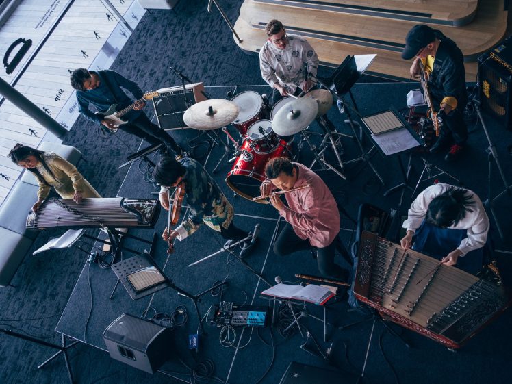 Image of A band of six musicians is performing indoors with various instruments, including drums, guitars, flutes, and traditional string instruments, seen from above. Sheet music and audio equipment are visible around them.