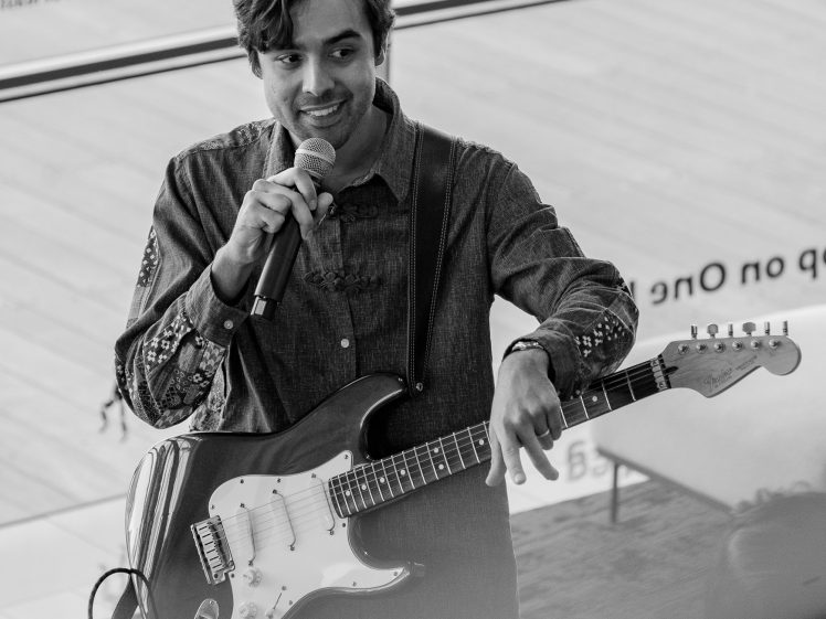 Image of A man holding a microphone and an electric guitar smiles whilst speaking, standing indoors near large windows. The image is in black and white.