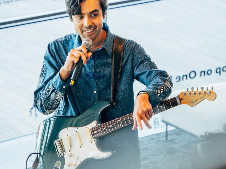 Image of A man wearing a blue denim shirt holds a microphone and electric guitar, smiling while speaking to an audience in a modern indoor space with large windows.