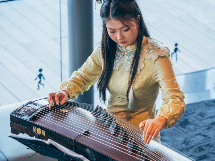 Image of A young woman in a yellow traditional dress plays a guzheng, a Chinese string instrument, indoors. She is focused on the instrument, with sheet music placed in front of her.