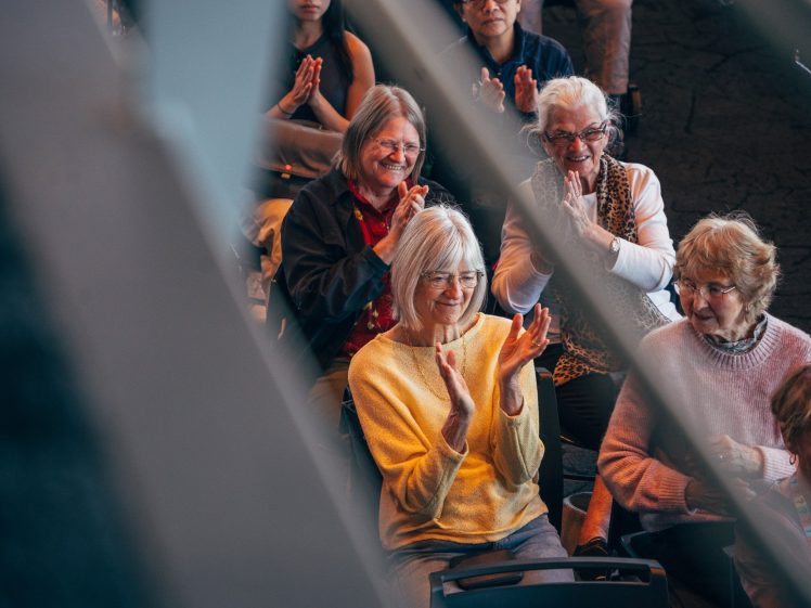 Image of A group of older women sit closely together, smiling and clapping, whilst enjoying an event. The image is taken through metal beams, partially obscuring the view.