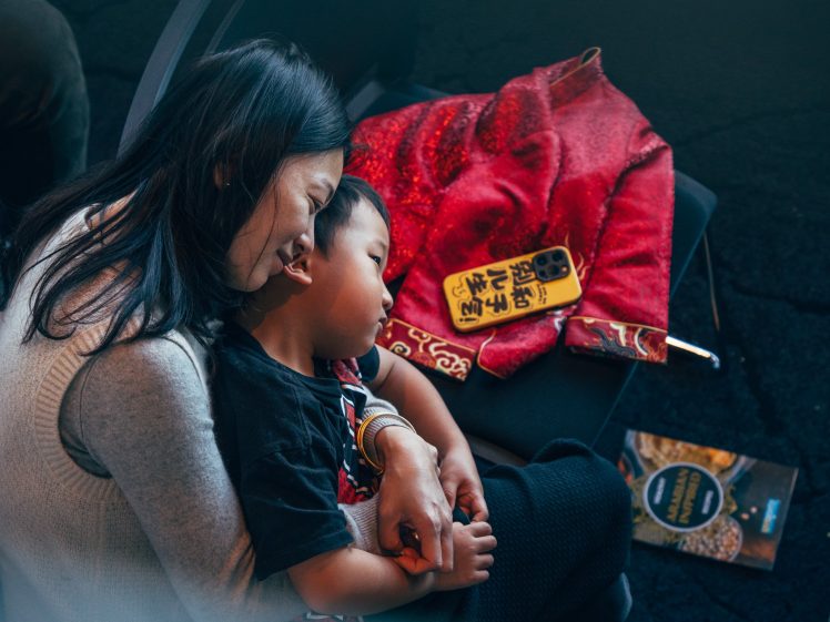 Image of A woman lovingly hugs a young boy who is resting on her lap. They are sitting on chairs with a red garment and a yellow item with Chinese writing nearby, and a book on the floor.