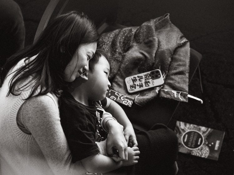 Image of A woman hugs a young child while sitting on a chair. They both smile and look relaxed. Nearby are a blanket, a phone with a decorated case, and a book on the floor. The photo is in black and white.