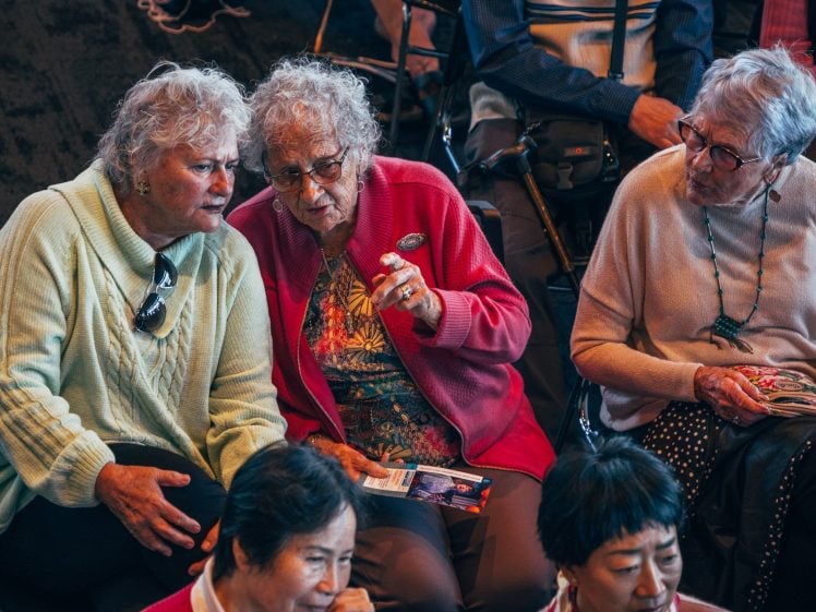 Image of Three elderly women sit closely together, engaged in conversation, whilst a group of people of various ages sit nearby. One woman gestures with her hand, and all appear attentive and sociable in a warm indoor setting.