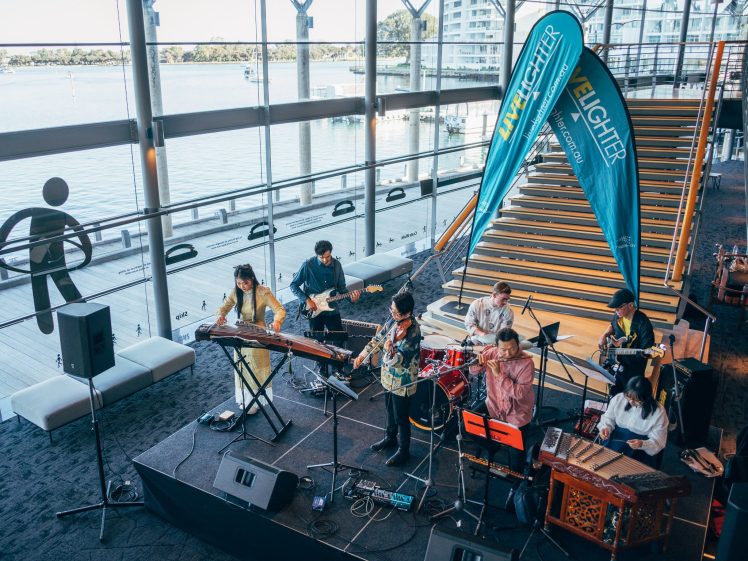 Image of A band performs on a small indoor stage by large windows overlooking water. Musicians play various instruments, including guitar, drums, and traditional instruments. A blue Live Lighter banner stands near a staircase.