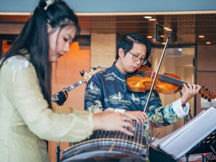 Image of Two musicians play traditional instruments indoors; a woman in the foreground plays a guzheng, whilst a man wearing glasses and patterned clothing in the background plays a violin. Music sheets are visible on a stand.