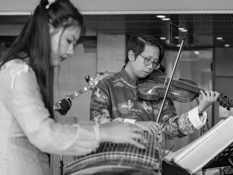 Image of A black and white photo of two musicians in traditional attire; a woman plays a stringed instrument in the foreground, and a man plays a violin in the background, both focussed on their performance.