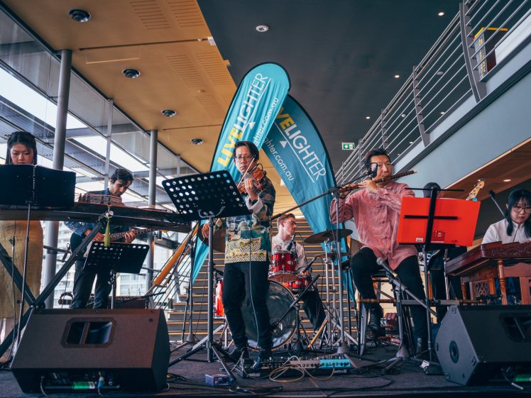 Image of A group of six musicians perform on stage indoors, playing various instruments including keyboard, guitar, violin, flute, and percussion. Banners and modern architecture are visible in the background.