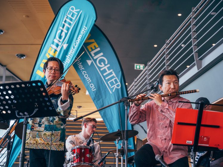Image of Three musicians perform indoors; one plays the violin, another plays the flute, and a third is on drums. Behind them are blue banners with VELIGHTER written on them and a modern staircase in the background.