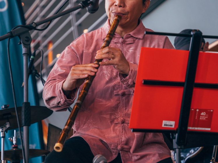 Image of A man in a pink shirt plays a bamboo flute on stage, seated behind a red music stand. Microphones and modern indoor railings are visible in the background.