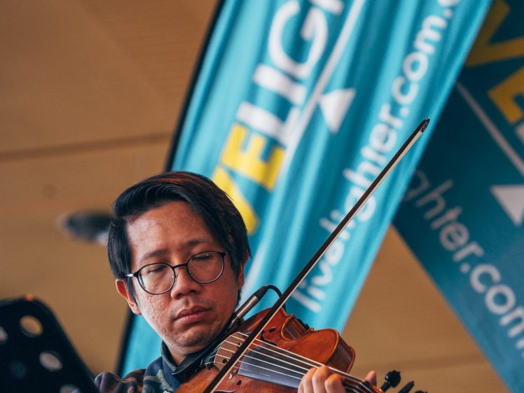 Image of A man wearing glasses plays a violin on stage; a blue banner in the background reads DELIGHTER and displays a web address. A music stand is visible in the foreground.