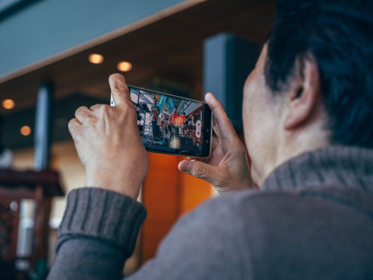 Image of A person wearing a brown jumper holds up a mobile phone, taking a photo or recording video indoors. The phone screen shows a group of people and colourful decorations. The image is taken from behind the person.