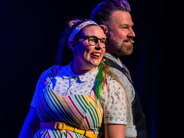 Image of Two performers stand back to back on stage, both smiling. The woman wears glasses, a colourful striped dress, and a headband, while the man has a beard and wears a waistcoat. Stage lighting highlights them against a dark background.
