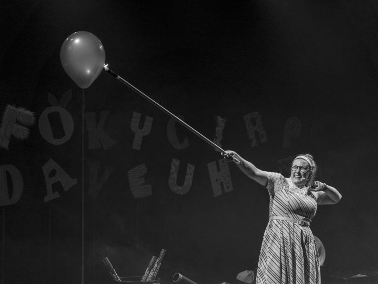Image of A woman in a striped dress smiles whilst holding a long stick with a balloon attached to the end on a stage. Large decorated letters hang in the background. The image is in black and white.