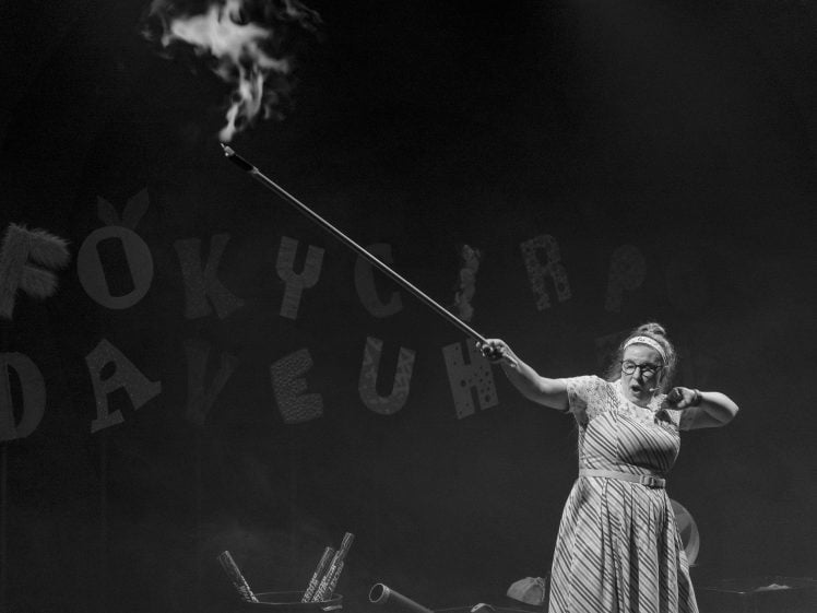 Image of A woman in a striped dress and glasses holds a long pole shooting out smoke on a dimly lit stage with jumbled letters hanging in the background.