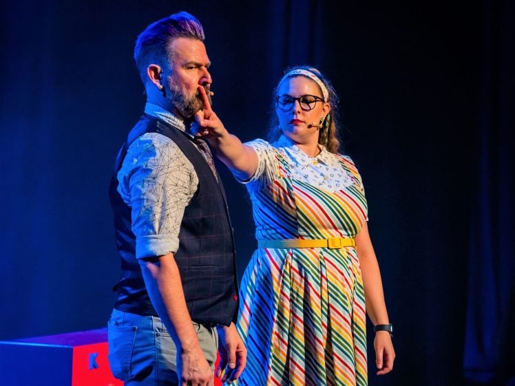 Image of A woman in a colourful striped dress holds her finger to the lips of a bearded man in a waistcoat and rolled-up sleeves on stage, both under dramatic, colourful lighting.