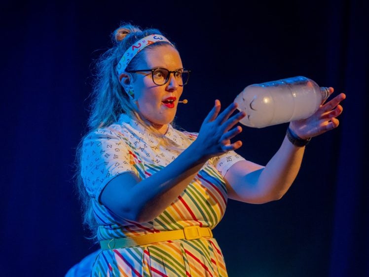 Image of A woman wearing glasses, a colourful striped dress, and a headband holds a clear plastic bottle filled with smoke or vapour on stage, appearing to perform a science demonstration.