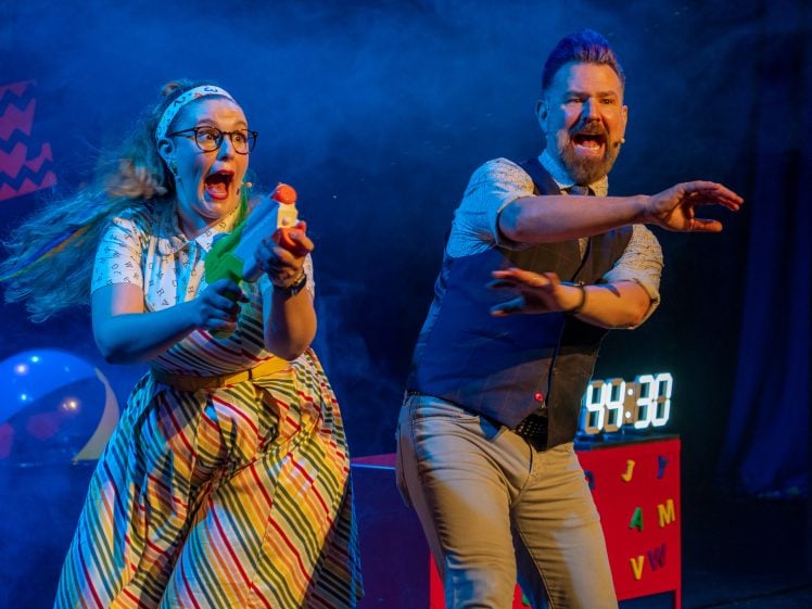 Image of Two performers on stage, one woman holding a water pistol and smiling excitedly, while a man beside her gestures forward; a digital timer and colourful props are visible in the background with dramatic stage lighting.