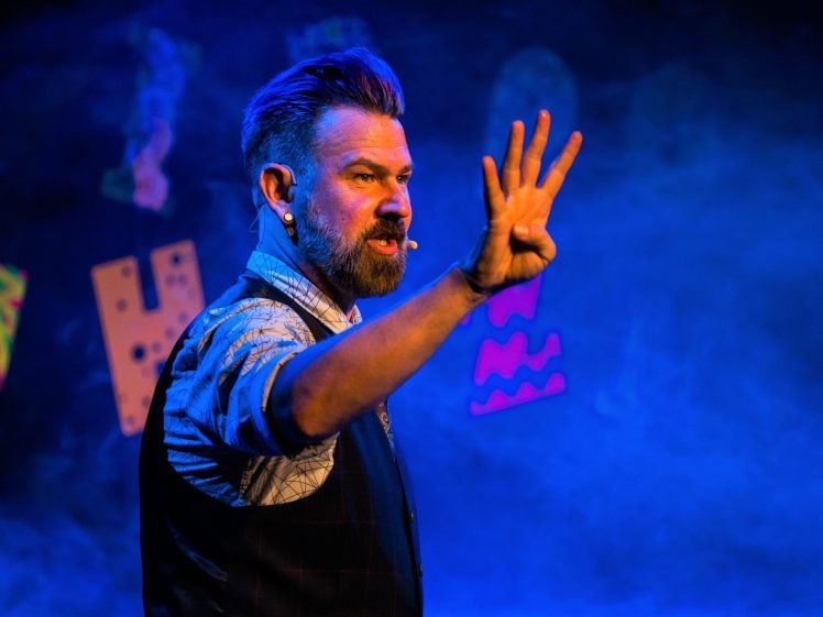 Image of A man with a beard and styled hair, wearing a waistcoat and headset, performs on stage under blue lighting, holding up four fingers. Colourful letters and shapes are visible in the background.