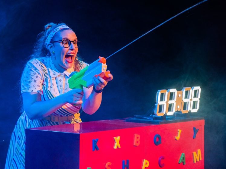 Image of A woman with glasses excitedly squirts water from a toy gun. She stands behind a red box decorated with colourful letters and a digital clock displaying 83:48. Blue lighting enhances the playful scene.