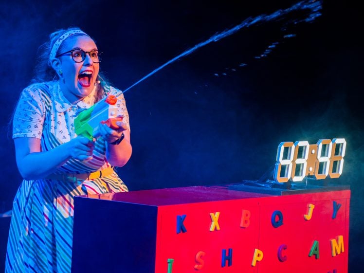 Image of A woman wearing glasses and a striped dress excitedly sprays water from a toy gun. She stands behind a red desk decorated with colourful letters and a digital clock displaying 88:88. Blue lighting adds a playful atmosphere.