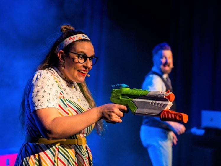 Image of A woman in a colourful striped dress and glasses smiles whilst holding a toy water pistol on stage, with a man blurred in the background. Both wear microphones, suggesting a performance or show.