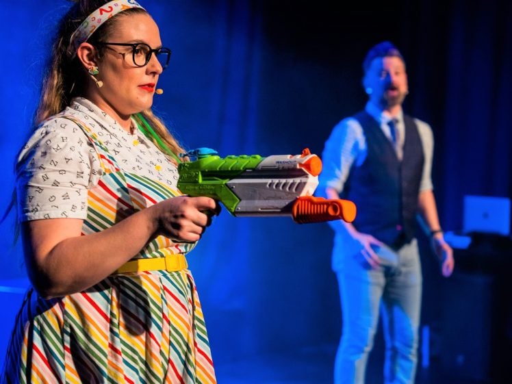 Image of A woman in glasses and a colourful striped dress aims a large toy water gun on stage, while a man in a waistcoat stands blurred in the background under stage lights.