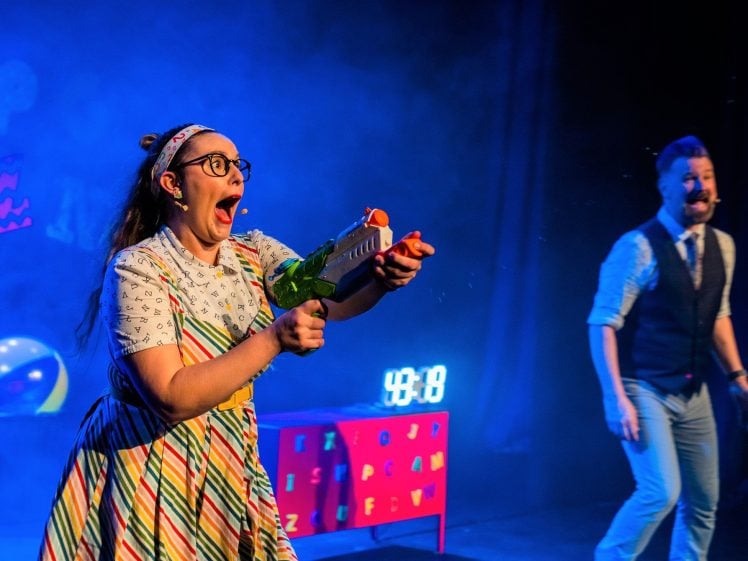 Image of A woman in colourful clothes and glasses excitedly sprays a water pistol onstage, whilst a man in a waistcoat and tie smiles in the background. The stage is decorated with bright props and has blue lighting.