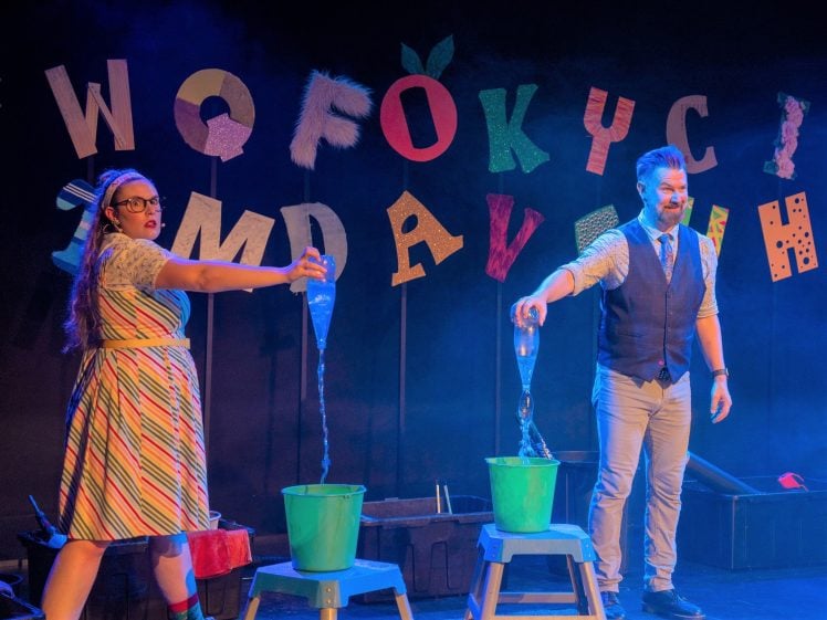 Image of Two performers stand on stage pouring water from plastic bottles into buckets on stools, with colourful letters on the wall behind them and blue stage lighting creating a playful atmosphere.