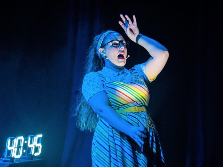 Image of A performer in a striped dress and headset sings or speaks passionately on stage. A digital clock reading “40:45” glows beside her in a dark, dramatic setting.